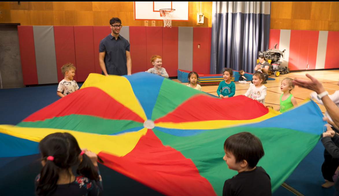 A group of children playing with a colorful kite in a gym.