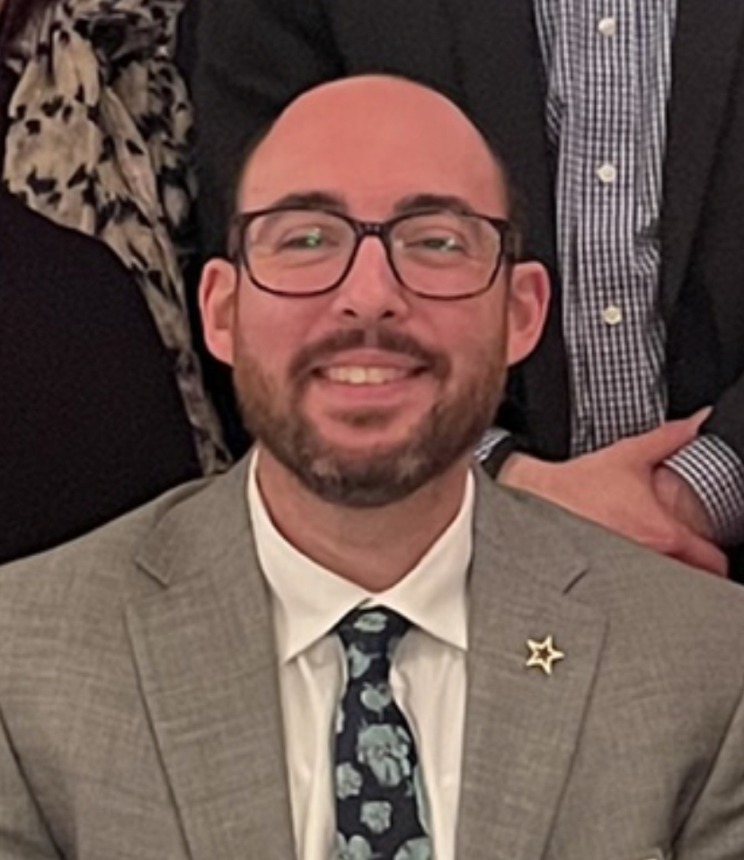 Jared Mintz, a man in a suit and tie, smiles in front of a group of people.