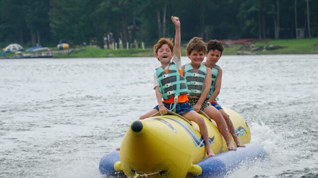Three boys at a summer camp riding on a banana boat on a lake.