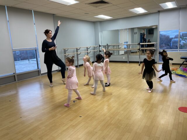 A dance instructor demonstrating a pose to a group of young girls in a ballet class during the March Dance Open House.