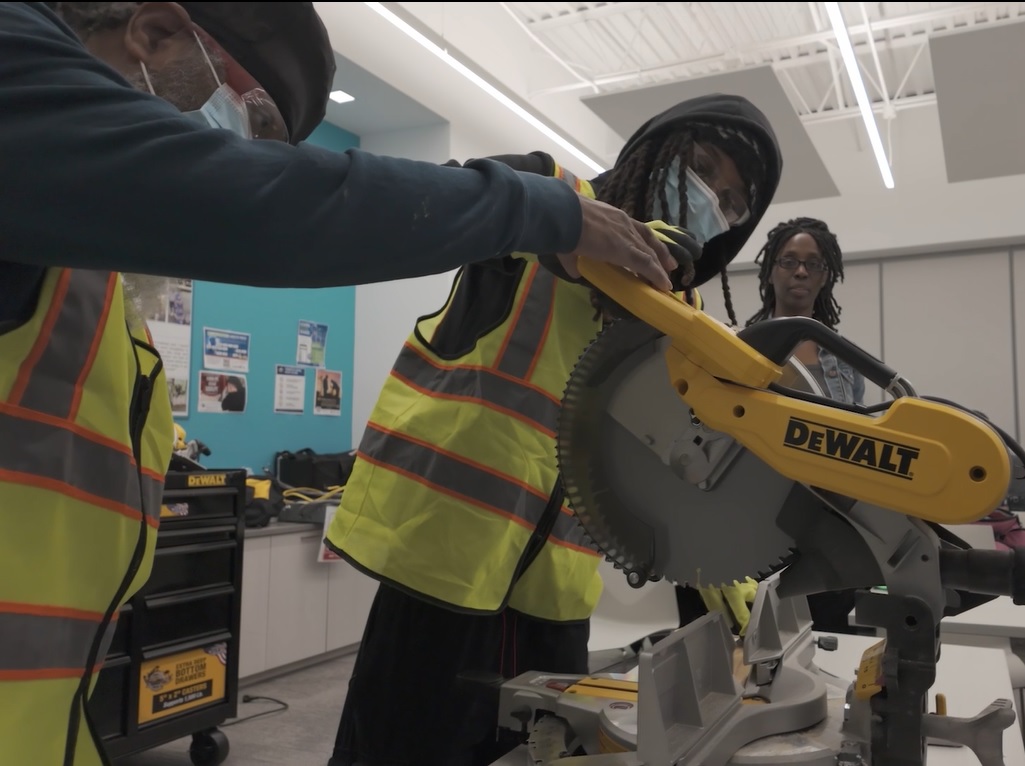 Two job seekers wearing safety vests and protective masks using a Dewalt miter saw in a workshop during construction skills training.