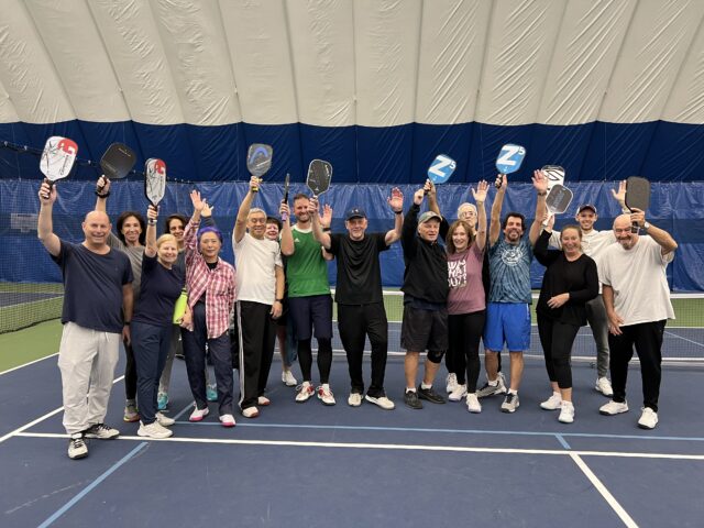 Group of people posing with pickleball paddles at an April Pickleball Clinic on an indoor court.