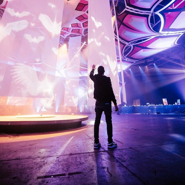 A person standing in a large hall with vibrant light projections and ornate patterns on towering structures, gesturing upwards during a cultural event.